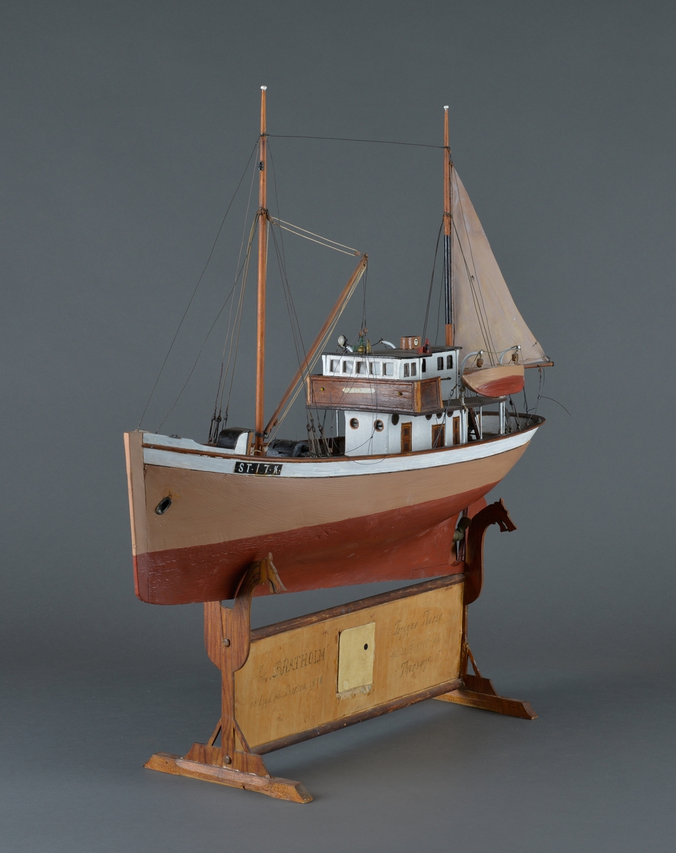 Fiskebåt eller sjark laget i trevirke. 
Båtmodellen står på et stativ laget av mørkt tre, antatt teak. Stativet består av ei plate med utskjæringer av drager og sjøfugl (terner?) Midt på platen er det felt inn et foto av en mann med blank-skjerm lue, kankskje produsenten(?). For at båtmodellen skal stå stødig, er det påmontert labber og utskårede dragehoder, slik at båten hviler i en slags krybbe. 
Båten har styrhus akter og nedgang til lugarer i baugen. 
Mast foran med bom og messanmast med gaffelsegl. To livbåter henger i daviter. 
Forseggjort og detaljert modellarbeid.