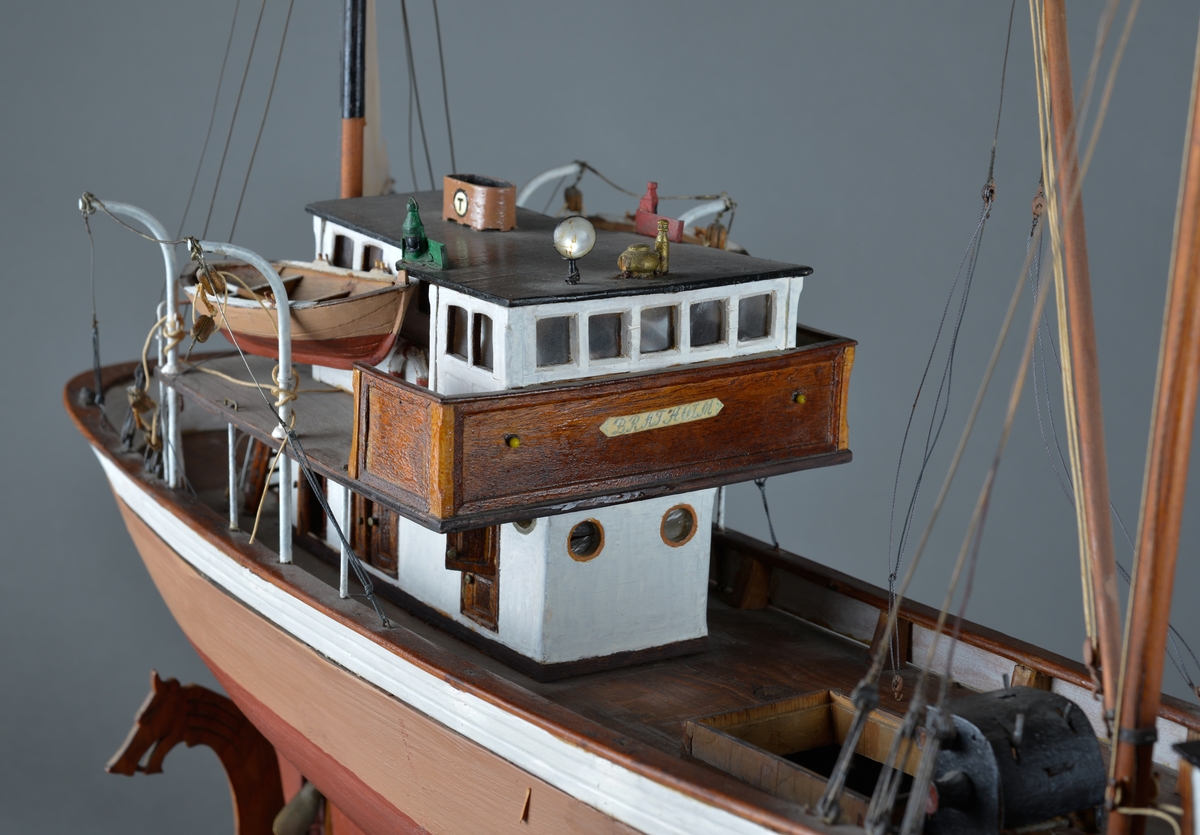 Fiskebåt eller sjark laget i trevirke. 
Båtmodellen står på et stativ laget av mørkt tre, antatt teak. Stativet består av ei plate med utskjæringer av drager og sjøfugl (terner?) Midt på platen er det felt inn et foto av en mann med blank-skjerm lue, kankskje produsenten(?). For at båtmodellen skal stå stødig, er det påmontert labber og utskårede dragehoder, slik at båten hviler i en slags krybbe. 
Båten har styrhus akter og nedgang til lugarer i baugen. 
Mast foran med bom og messanmast med gaffelsegl. To livbåter henger i daviter. 
Forseggjort og detaljert modellarbeid.