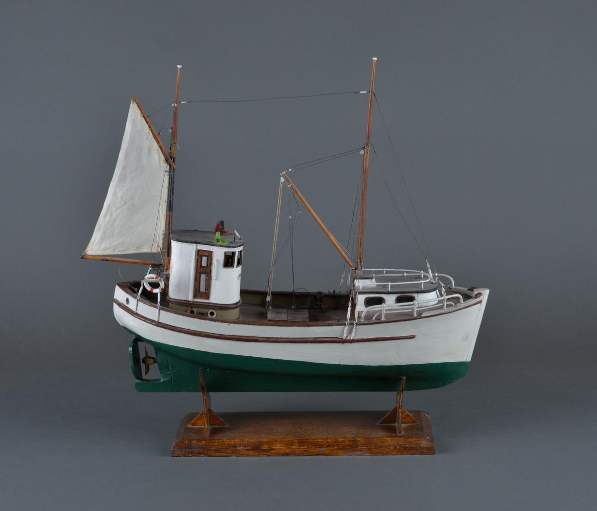 Fiskebåt eller sjark laget i trevirke. Båtmodellen står på et stativ laget av mørkt tre, antatt teak. Støttene har utskjæringer. 
Båten har styrhus, nedgang til lugarene i baugen. Mast foran med bom og messanmast med gaffelsegl.