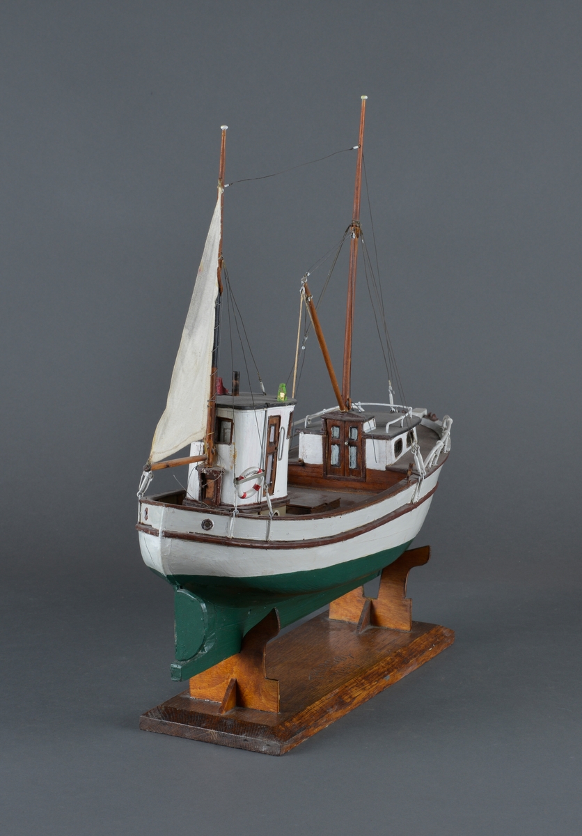 Fiskebåt eller sjark laget i trevirke. Båtmodellen står på et stativ laget av mørkt tre, antatt teak. Støttene har utskjæringer. 
Båten har styrhus, nedgang til lugarene i baugen. Mast foran med bom og messanmast med gaffelsegl.