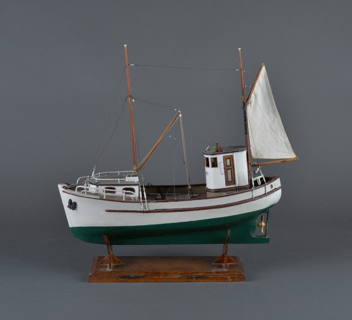 Fiskebåt eller sjark laget i trevirke. Båtmodellen står på et stativ laget av mørkt tre, antatt teak. Støttene har utskjæringer. 
Båten har styrhus, nedgang til lugarene i baugen. Mast foran med bom og messanmast med gaffelsegl.