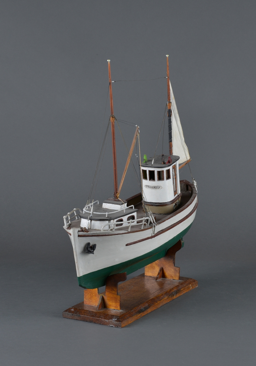 Fiskebåt eller sjark laget i trevirke. Båtmodellen står på et stativ laget av mørkt tre, antatt teak. Støttene har utskjæringer. 
Båten har styrhus, nedgang til lugarene i baugen. Mast foran med bom og messanmast med gaffelsegl.