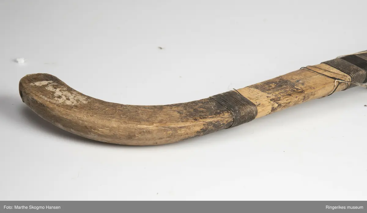 Hockeykølle i tre med rundt skaft og  kølle/slagdel som er flat på den ene siden og rund på den andre. Skaftet er dekket med tråd i to farger som er surret tett rundt skaftet.  