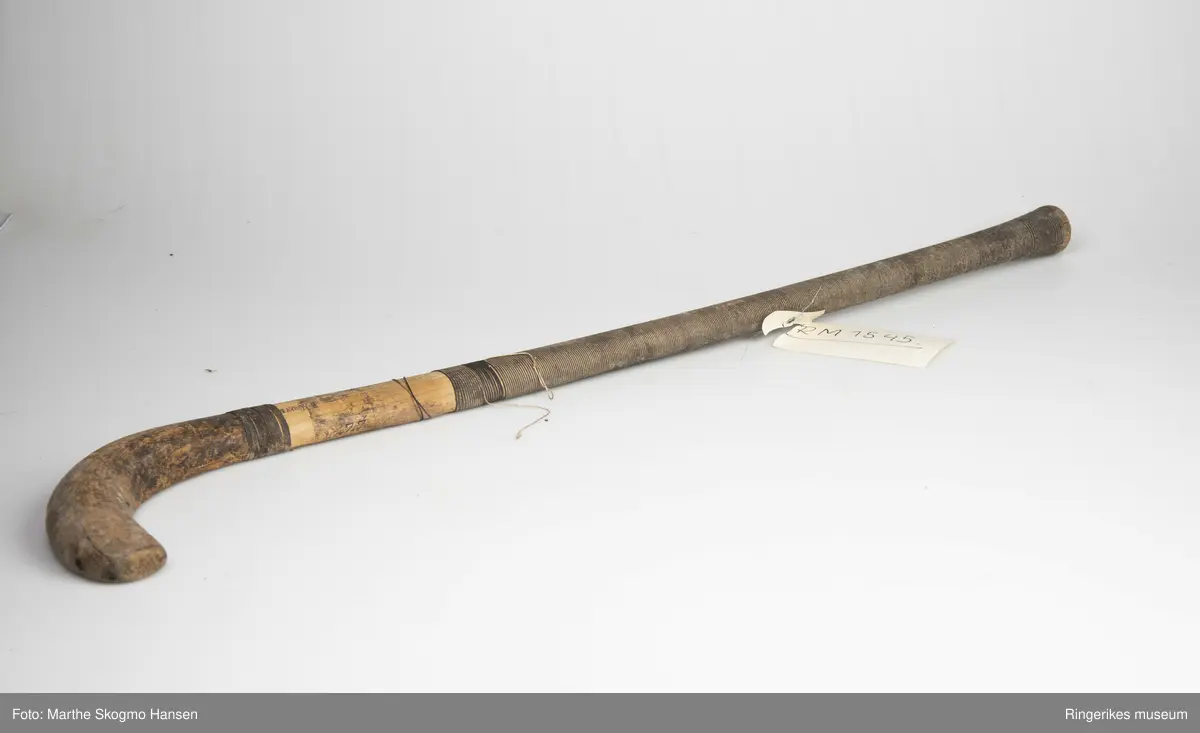 Hockeykølle i tre med rundt skaft og  kølle/slagdel som er flat på den ene siden og rund på den andre. Skaftet er dekket med tråd i to farger som er surret tett rundt skaftet.  
