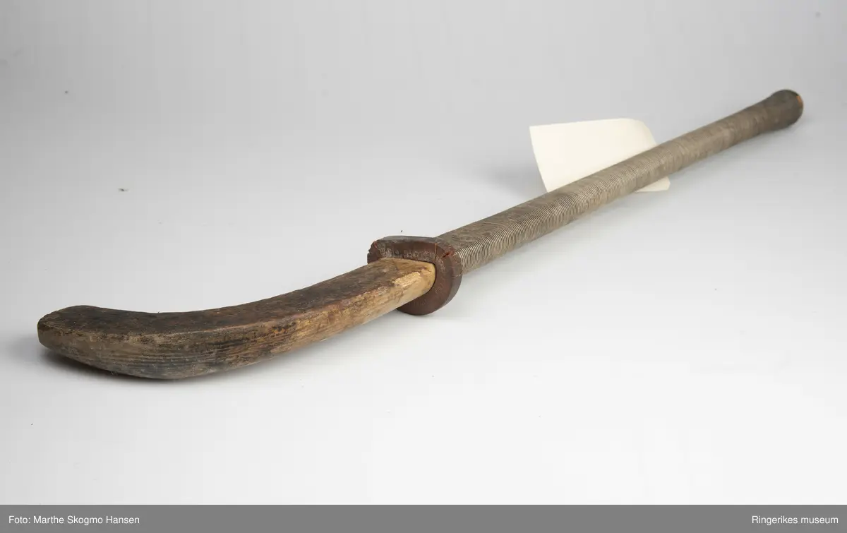 Hockeykølle i tre med rundt skaft og  kølle/slagdel som er flat på den ene siden og rund på den andre.Skaftet er dekket med tråd i to farger som er surret tett rundt skaftet.  I overgangen mellom skaft og kølle er det en rød gummiring.