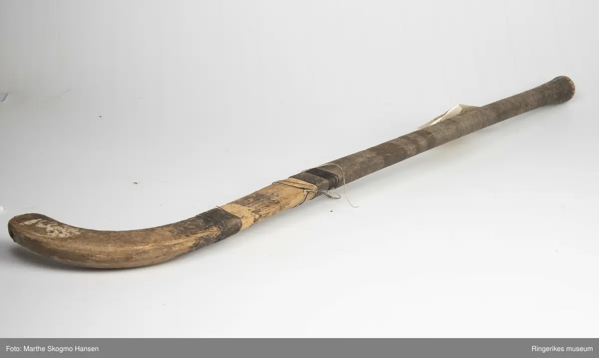 Hockeykølle i tre med rundt skaft og  kølle/slagdel som er flat på den ene siden og rund på den andre. Skaftet er dekket med tråd i to farger som er surret tett rundt skaftet.  