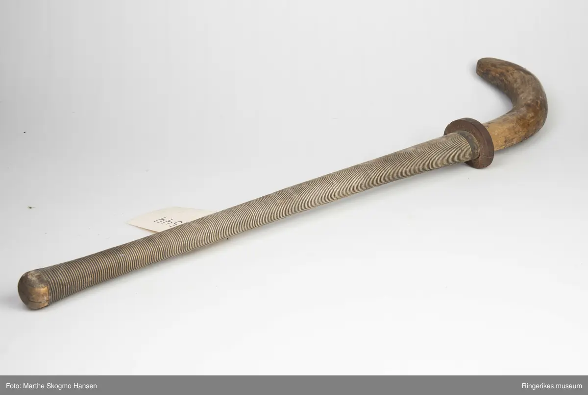 Hockeykølle i tre med rundt skaft og  kølle/slagdel som er flat på den ene siden og rund på den andre.Skaftet er dekket med tråd i to farger som er surret tett rundt skaftet.  I overgangen mellom skaft og kølle er det en rød gummiring.