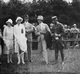 Gruppfoto; ett sällskap om fem personer står på ett fält, 1920-tal. I bakgrunden står en folksamling. Två civila kvinnor, två civila män och en flygofficer. Eventuellt i samband med flygdag / flyguppvisning.