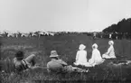 Ett sällskap om fem personer sitter på ett flygfält vid en flygdag, 1920-tal. I bakgrunden står militära flygplan samt en folksamling.