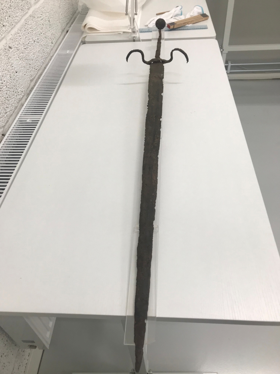 Dubbeleggat, närmast komplett svärd. Avslutas med en rund svärdskanpp som nu är löst fäst vid greppets tånge. Parerstången är böjd i två bågar.