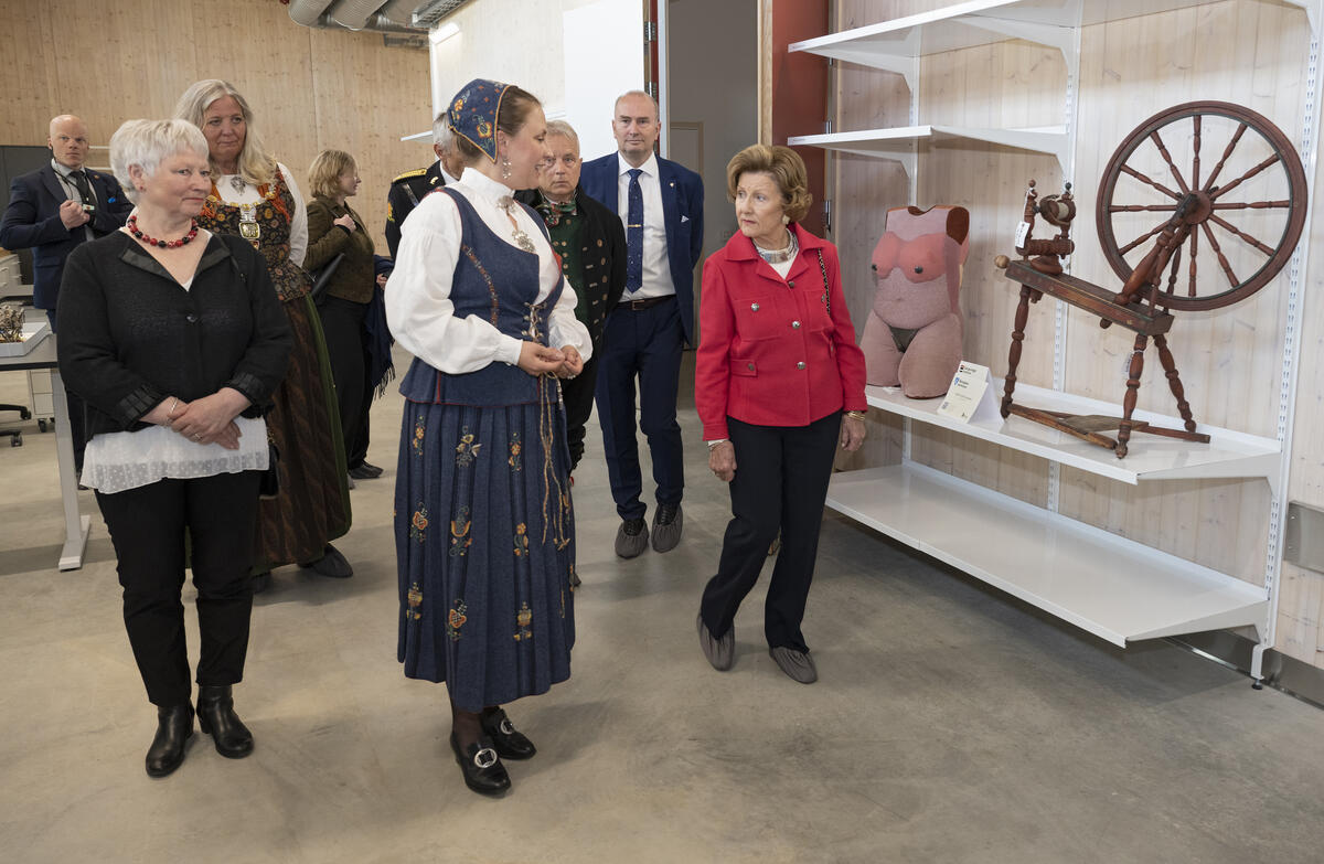 En gruppe personer i dresser, bunader og pene klær. først i gruppa snakker to av personene samen, og til høyre for dem står en gammel spinnerokke og mannekeng