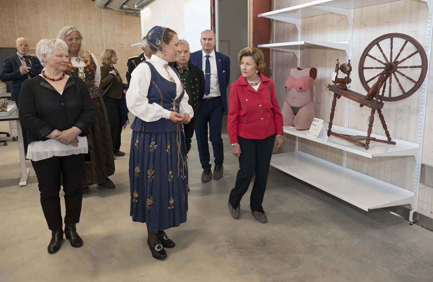 En gruppe personer i dresser, bunader og pene klær. først i gruppa snakker to av personene samen, og til høyre for dem står en gammel spinnerokke og mannekeng