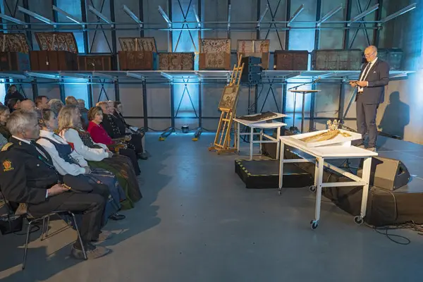 En mann i dress som holder tale for en gruppe personer i seremonielle eller pene klær foran han, som er til venstre taleren i bilde