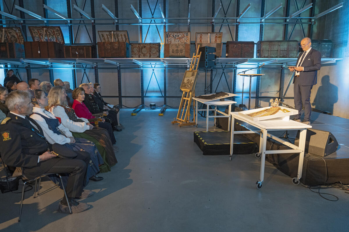 En mann i dress som holder tale for en gruppe personer i seremonielle eller pene klær foran han, som er til venstre taleren i bilde