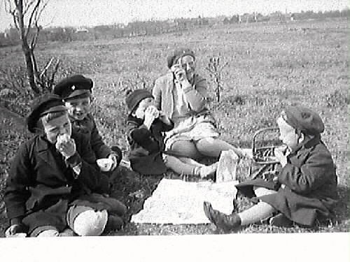 Utflykt. "Några av barnen med sköterska på utflykt." Fyra små barnhemsbarn har picnic med en ung sköterska på en äng.