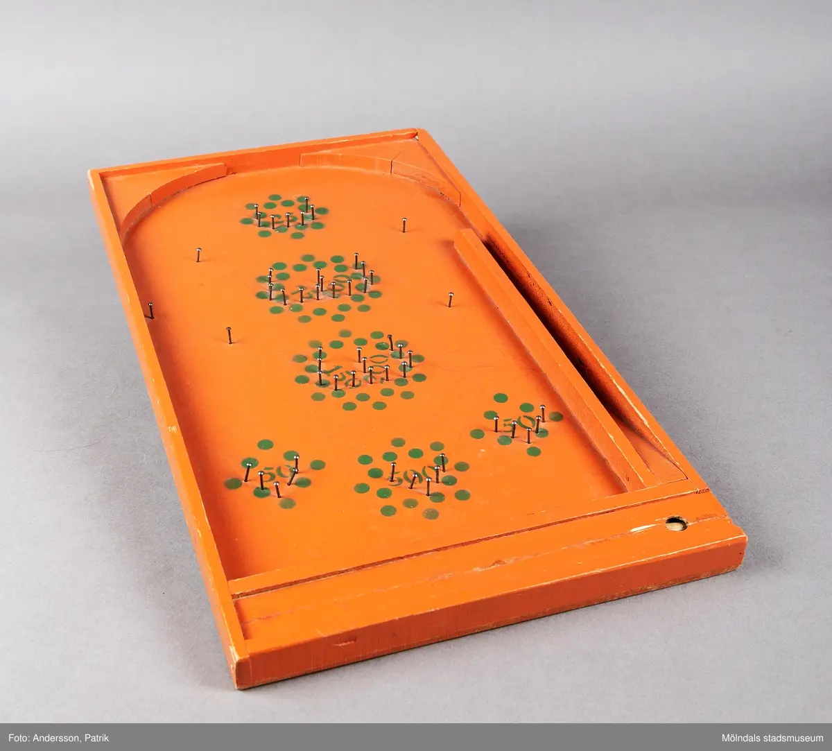 Fortunaspel bestående av en bottenplatta av bemålad masonit med kanter och ett fack av bemålat trä.
I facket finns en kort kö, två fotpinnar och metallkulor. Planen är försedd med spikar och en grön dekor som anger värdet. På vänster ytterkant är initialerna E.L skrivna med blyerts.
Spelet är handgjort och målat med orange färg. Punkter och siffror är målade i grön färg. Längst ned på spelet fins ett litet fack med speltillbehör. I facket finns en kort kö, två fotpinnar och metallkulor. Facket har ett lock som kan skjutas in. Spelet är förmodligen tillverkat på 50-talet och är en föregångare till flipperspelet. Poäng uppnås genom att kulor skjuts iväg med en pinne  och därefter hamnar i fack med olika värden.