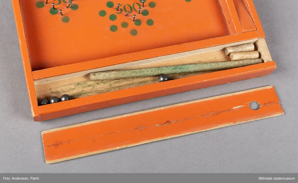 Fortunaspel bestående av en bottenplatta av bemålad masonit med kanter och ett fack av bemålat trä.
I facket finns en kort kö, två fotpinnar och metallkulor. Planen är försedd med spikar och en grön dekor som anger värdet. På vänster ytterkant är initialerna E.L skrivna med blyerts.
Spelet är handgjort och målat med orange färg. Punkter och siffror är målade i grön färg. Längst ned på spelet fins ett litet fack med speltillbehör. I facket finns en kort kö, två fotpinnar och metallkulor. Facket har ett lock som kan skjutas in. Spelet är förmodligen tillverkat på 50-talet och är en föregångare till flipperspelet. Poäng uppnås genom att kulor skjuts iväg med en pinne  och därefter hamnar i fack med olika värden.