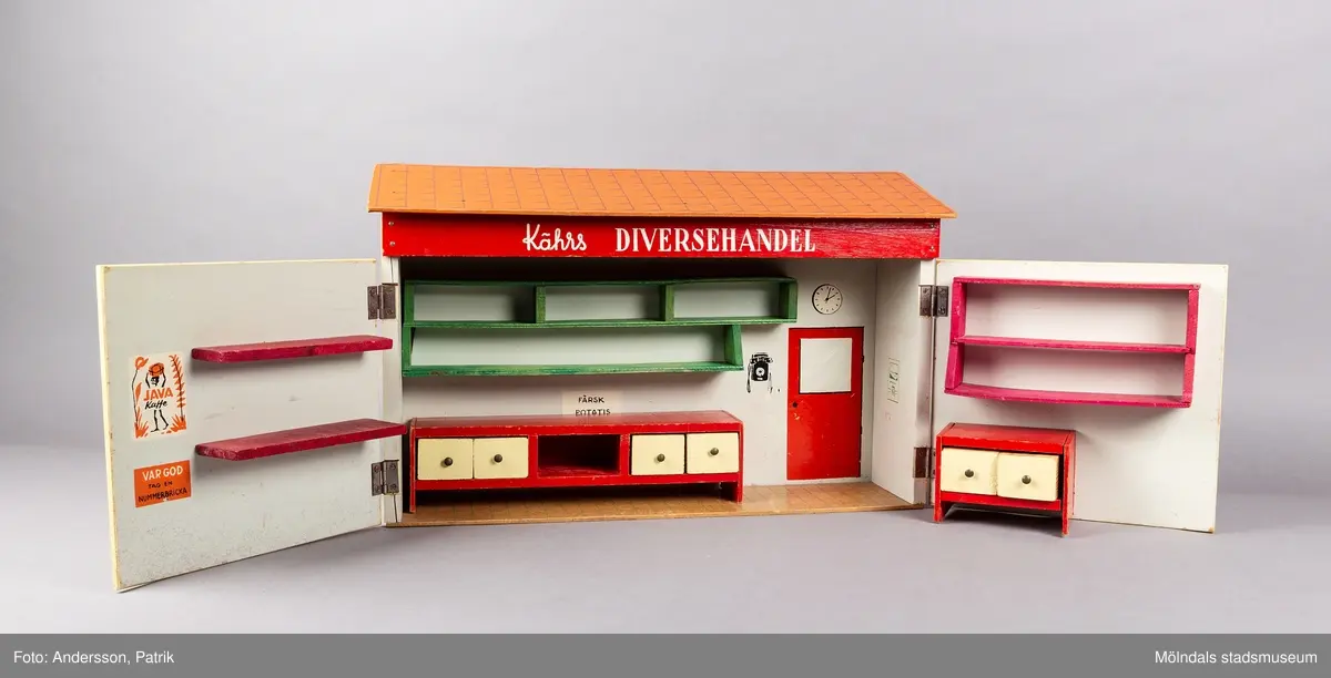 Leksak i trä i form av en liten affär. På framsidan finns texten "Kährs Diversehandel", dekor i form av två skyltfönster och en liten dörr med ett handtag i metall. Fronten består av två luckor går att öppna. Inuti finns en butiksinredning med gröna hyllor och en lös röd butiksdisk med sex gula utdragbara lådor.
Allt är tillverkat i trä. Affärens utsida är målad i en ljusgul färg, med ett tak som är bemålat med tegelröda tegelpannor. Under takåsen löper en röd list med texten "Kährs Diversehandel" målat med vita bokstäver.
Leksaksbutiken är tillverkad av Kährs. Företaget grundades 1857 och tillverkade till en början leksaker och möbler, men tillverkningen övergick efter andra världskriget alltmer till att omfatta endast dörrar och golv.