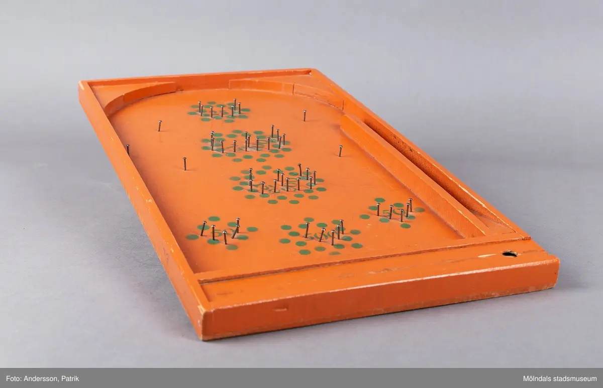Fortunaspel bestående av en bottenplatta av bemålad masonit med kanter och ett fack av bemålat trä.
I facket finns en kort kö, två fotpinnar och metallkulor. Planen är försedd med spikar och en grön dekor som anger värdet. På vänster ytterkant är initialerna E.L skrivna med blyerts.
Spelet är handgjort och målat med orange färg. Punkter och siffror är målade i grön färg. Längst ned på spelet fins ett litet fack med speltillbehör. I facket finns en kort kö, två fotpinnar och metallkulor. Facket har ett lock som kan skjutas in. Spelet är förmodligen tillverkat på 50-talet och är en föregångare till flipperspelet. Poäng uppnås genom att kulor skjuts iväg med en pinne  och därefter hamnar i fack med olika värden.