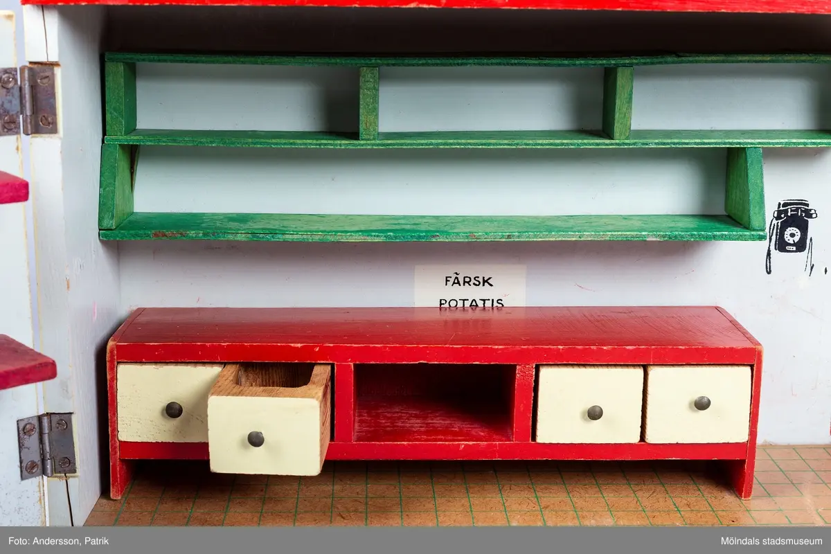 Leksak i trä i form av en liten affär. På framsidan finns texten "Kährs Diversehandel", dekor i form av två skyltfönster och en liten dörr med ett handtag i metall. Fronten består av två luckor går att öppna. Inuti finns en butiksinredning med gröna hyllor och en lös röd butiksdisk med sex gula utdragbara lådor.
Allt är tillverkat i trä. Affärens utsida är målad i en ljusgul färg, med ett tak som är bemålat med tegelröda tegelpannor. Under takåsen löper en röd list med texten "Kährs Diversehandel" målat med vita bokstäver.
Leksaksbutiken är tillverkad av Kährs. Företaget grundades 1857 och tillverkade till en början leksaker och möbler, men tillverkningen övergick efter andra världskriget alltmer till att omfatta endast dörrar och golv.