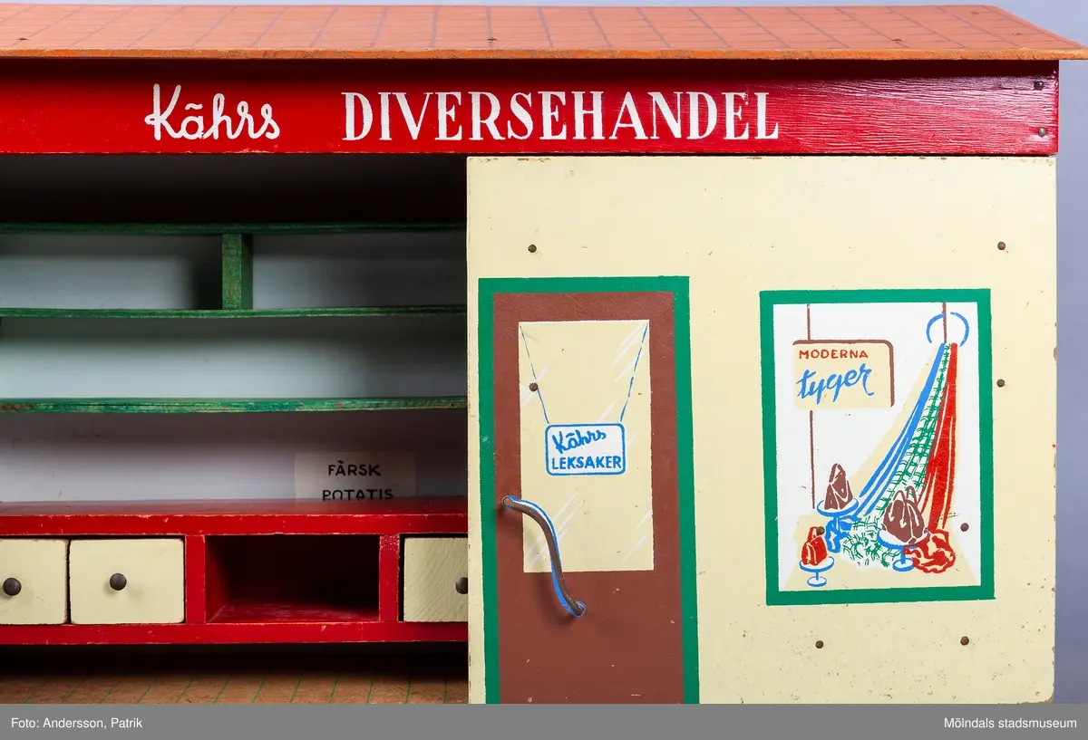 Leksak i trä i form av en liten affär. På framsidan finns texten "Kährs Diversehandel", dekor i form av två skyltfönster och en liten dörr med ett handtag i metall. Fronten består av två luckor går att öppna. Inuti finns en butiksinredning med gröna hyllor och en lös röd butiksdisk med sex gula utdragbara lådor.
Allt är tillverkat i trä. Affärens utsida är målad i en ljusgul färg, med ett tak som är bemålat med tegelröda tegelpannor. Under takåsen löper en röd list med texten "Kährs Diversehandel" målat med vita bokstäver.
Leksaksbutiken är tillverkad av Kährs. Företaget grundades 1857 och tillverkade till en början leksaker och möbler, men tillverkningen övergick efter andra världskriget alltmer till att omfatta endast dörrar och golv.
