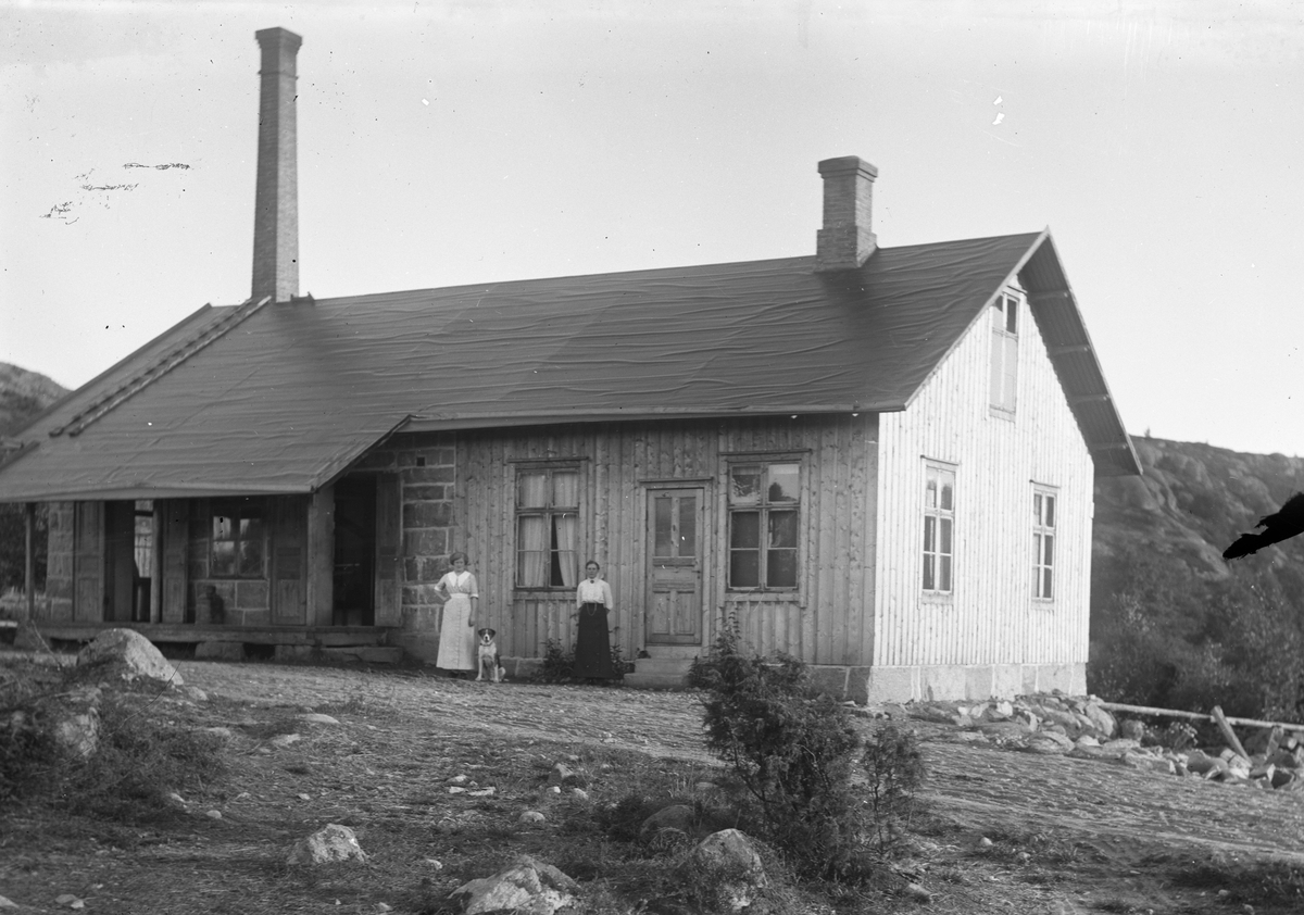 Två kvinnor, troligen mejerskor, står med hunden Nobis vid Mejeriet i Grimmared. Byggnaden har blottad stenstomme vid lastbryggan men träfasad i övrigt.