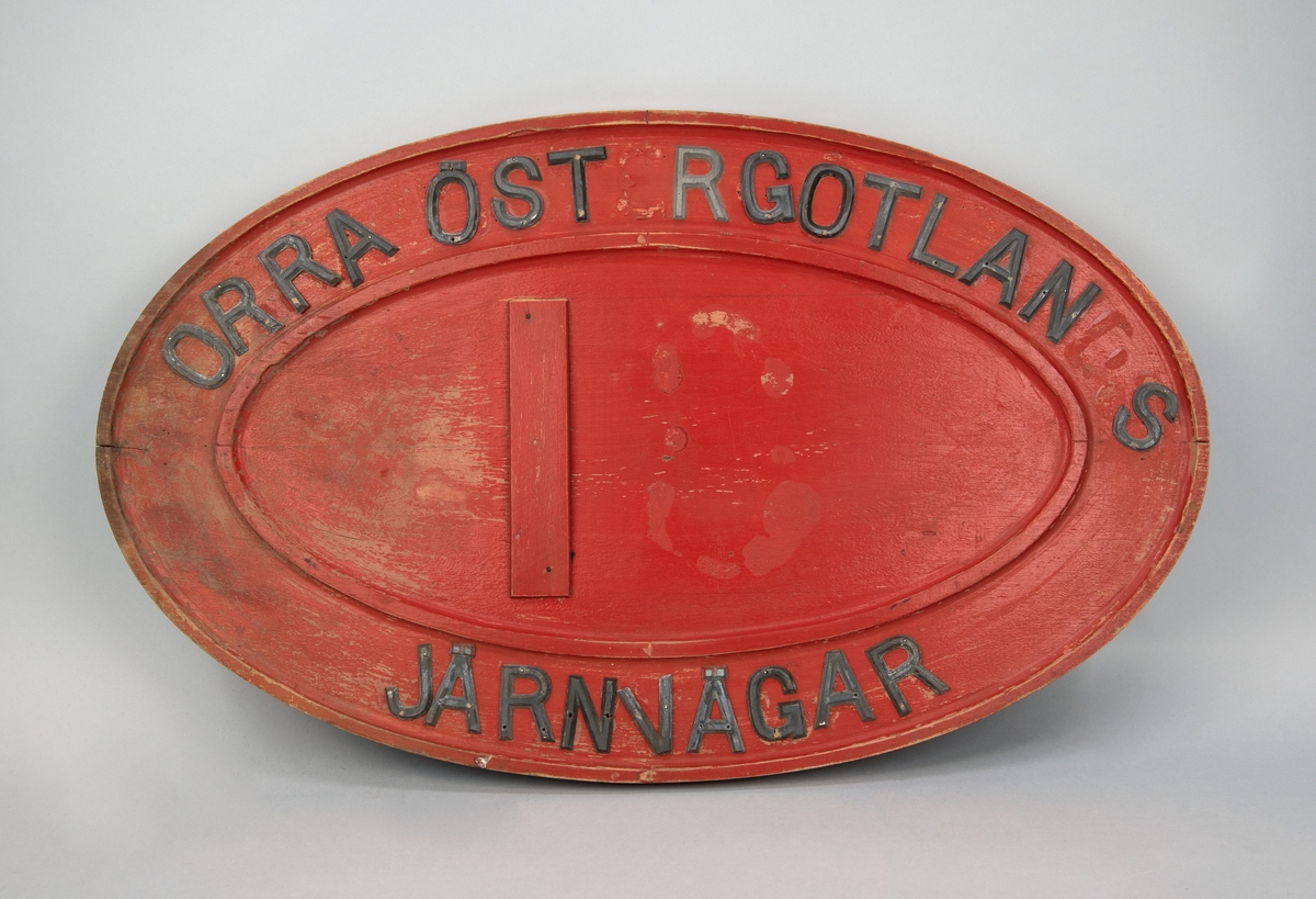 Oval gjutförlaga till loknummerskylt av rödmålat trä. Med fastspikade bokstäver av metall står det upptill  "ORRA ÖST RGOTLAN S", nertill "JÄRNVÄGAR", i mitten "1" samt rester av limm i form av "8".