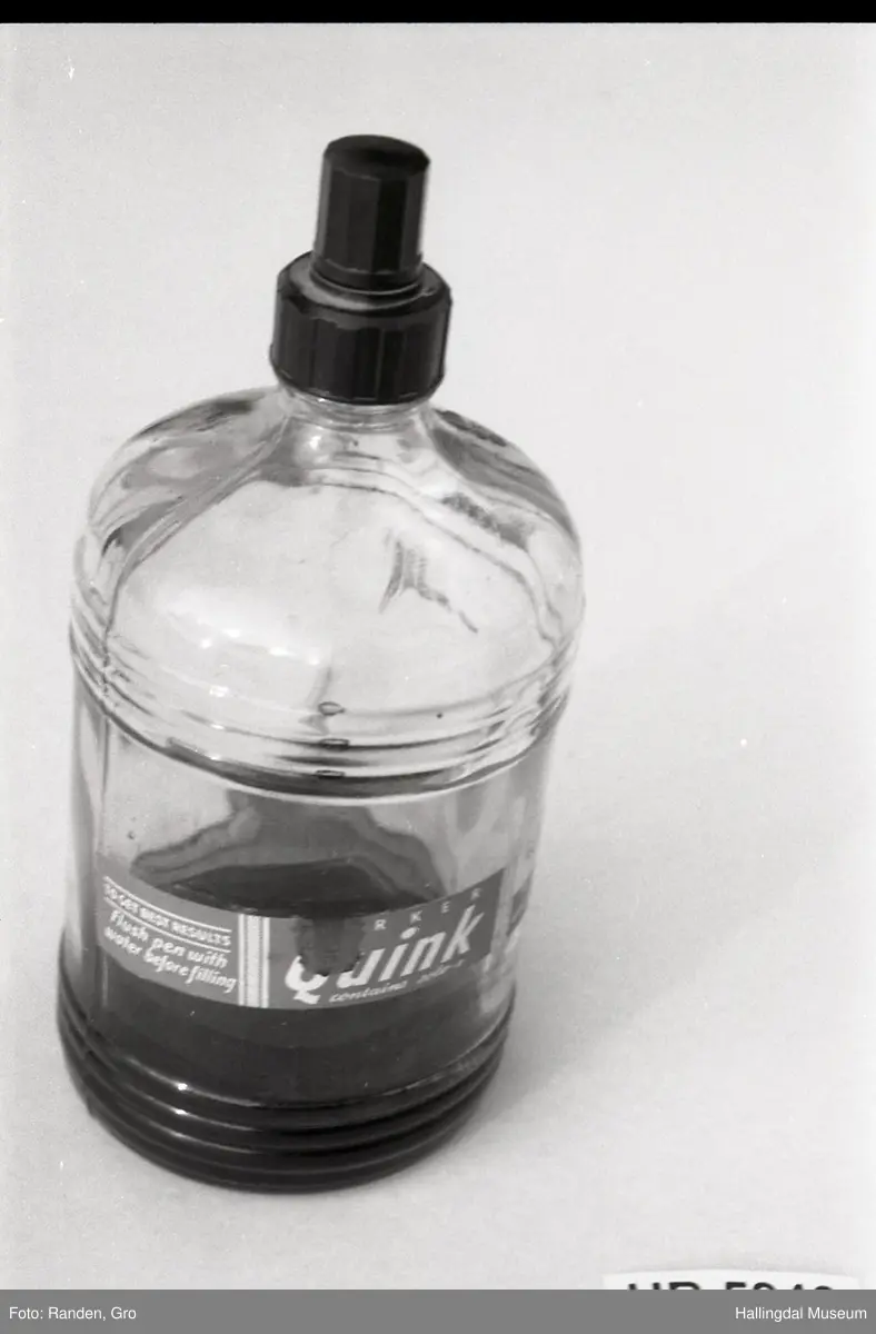 Flaske av blankt glas. Rett, sirkulært tverrsnitt. Det er litt blekk på flaska. Skruvekork av svart plast.