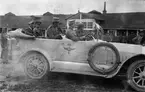 En samling militärer åker i Flygkompaniets bil, omkring 1920. Vy från sidan. I bakgrunden syns byggnader och en folksamling.