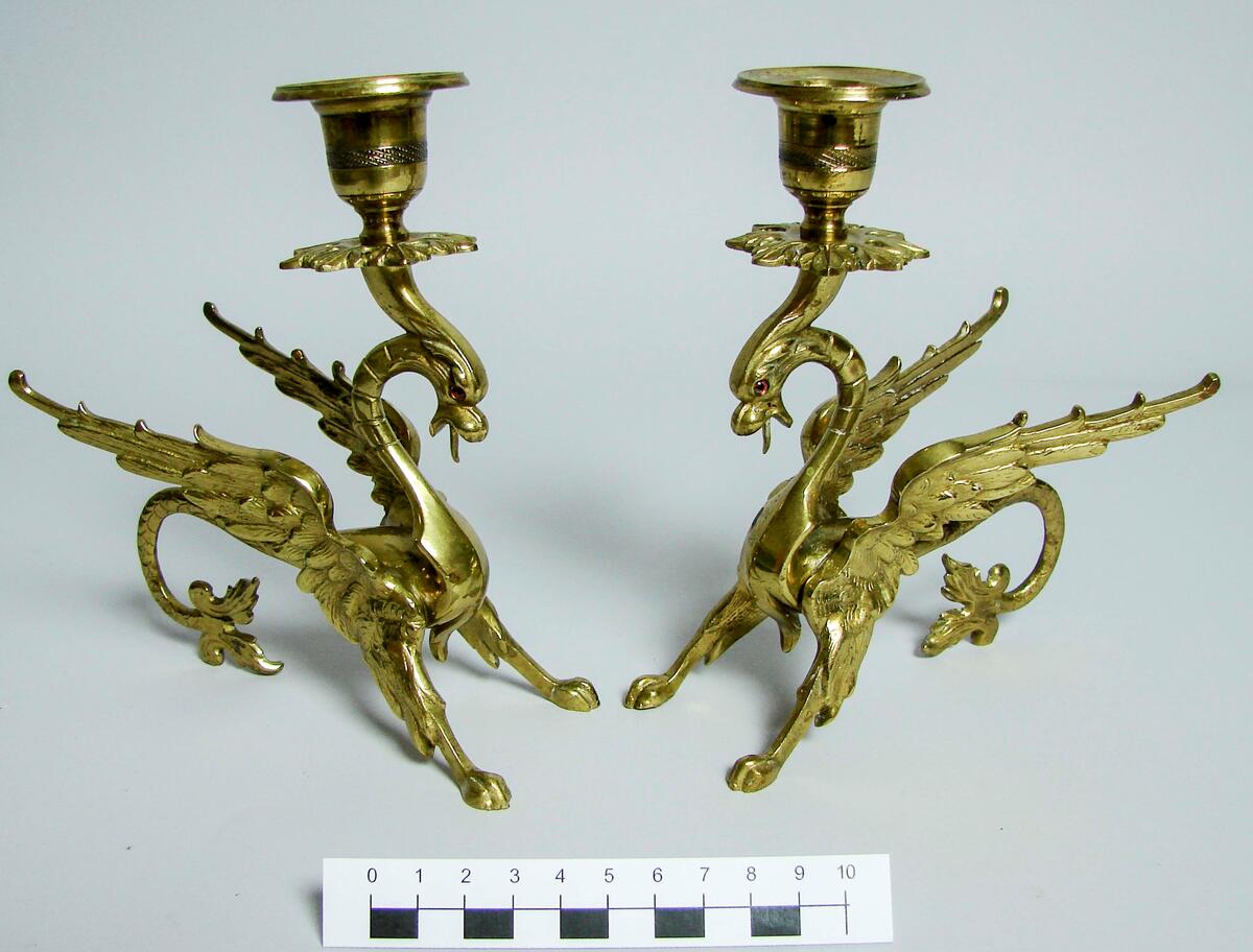 Ett par ljusstakar av mässing, ljushållare för 1 ljus vilar på huvudet av ett fantasidjur med drakhuvud och svans, rovfågelsvingar och ben samt lejontassar. Ögon av rött glas. Gåva: Länsassessor Tage Ljunggren, Härnösand 1961. (tillh. Fadern kassör Simon L.)
