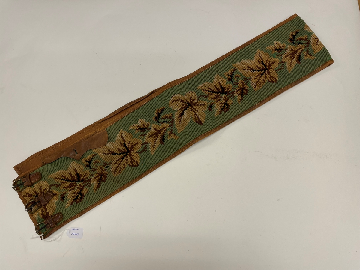 Pälsskärp, fodrat med ljusbrunt skinn. Broderat med korsstygn, grön botten, blad i flera färger, brunt.