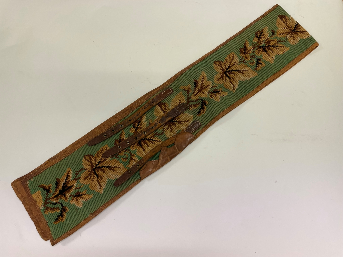 Pälsskärp, fodrat med ljusbrunt skinn. Broderat med korsstygn, grön botten, blad i flera färger, brunt.