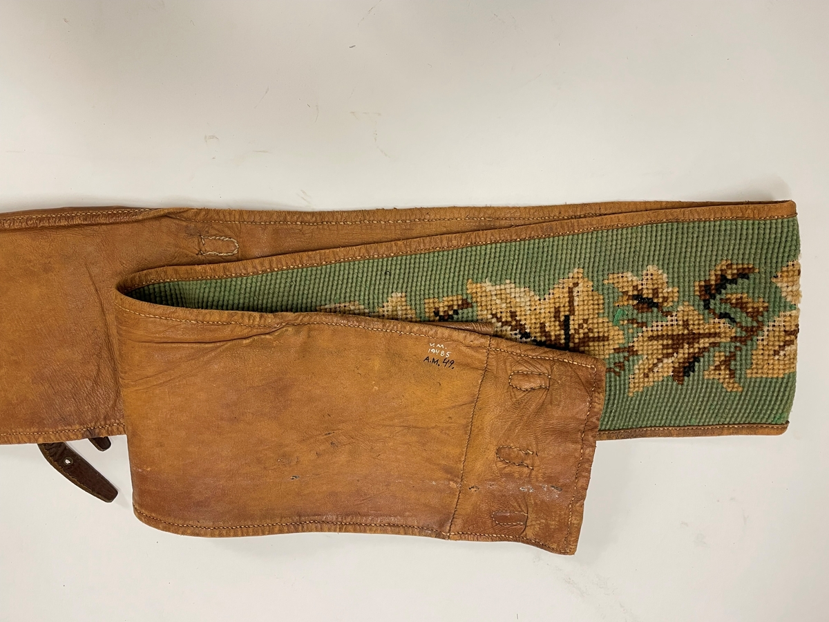 Pälsskärp, fodrat med ljusbrunt skinn. Broderat med korsstygn, grön botten, blad i flera färger, brunt.