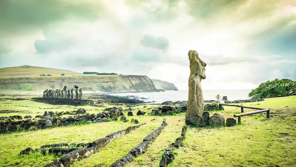 Vi ser et grønt landskap som ser ut til å ligge ved en klippe. Sjøen er rett nedenfor. Foran i bildet står det en oppreist figur i stein