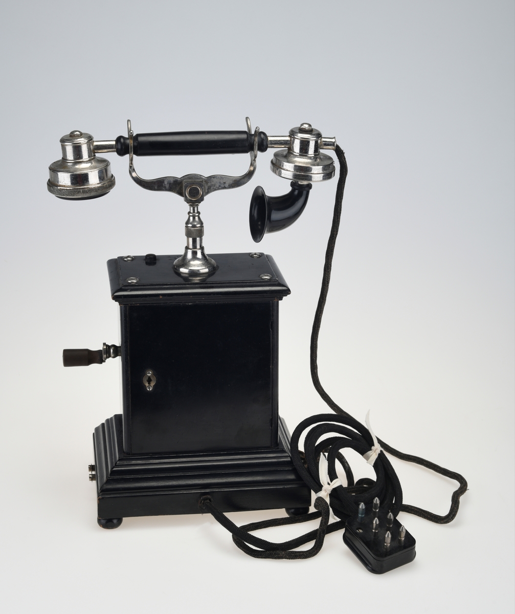 En bordtelefon av gammel modell laget av metall, tre og bakelitt. Foten på kassen har en ramme av svartlakkert metall og står på fire runde svartmalte trebein. På rammen er det montert på profilert treverk som også er svartmalt. Telefonkassen er av svart metall med en treplate med profilering på toppen som er skrudd fast. På midten er det en gaffel av sølvfarget metall der telefonrøret skal ligg. På den ene siden av kassen er det en sveiv av sølvfarget metall og håndtak av brun bakelitt. Ved siden av gaffelen er det en liten knapp som kan trykkes ned. Håndtaket på telefonrøret er av svart bakelitt. Høre- og snakkedelen er laget av blankt metall. På høredelen er det en rund bakelittplate med 7 hull. På snakkedelen er det et horn av svart bakelitt (har form som et lite grammofonhorn). Nede på den ene kortsiden går ledningen til telefonen ut. Den er lang og ender i et støpsel av svart bakelitt med seks metallplugger.