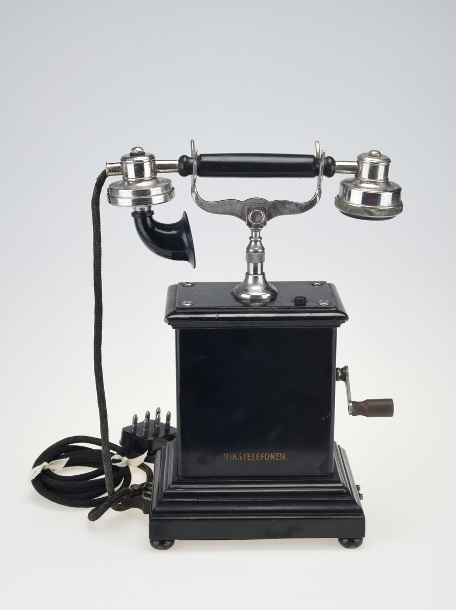 En bordtelefon av gammel modell laget av metall, tre og bakelitt. Foten på kassen har en ramme av svartlakkert metall og står på fire runde svartmalte trebein. På rammen er det montert på profilert treverk som også er svartmalt. Telefonkassen er av svart metall med en treplate med profilering på toppen som er skrudd fast. På midten er det en gaffel av sølvfarget metall der telefonrøret skal ligg. På den ene siden av kassen er det en sveiv av sølvfarget metall og håndtak av brun bakelitt. Ved siden av gaffelen er det en liten knapp som kan trykkes ned. Håndtaket på telefonrøret er av svart bakelitt. Høre- og snakkedelen er laget av blankt metall. På høredelen er det en rund bakelittplate med 7 hull. På snakkedelen er det et horn av svart bakelitt (har form som et lite grammofonhorn). Nede på den ene kortsiden går ledningen til telefonen ut. Den er lang og ender i et støpsel av svart bakelitt med seks metallplugger.