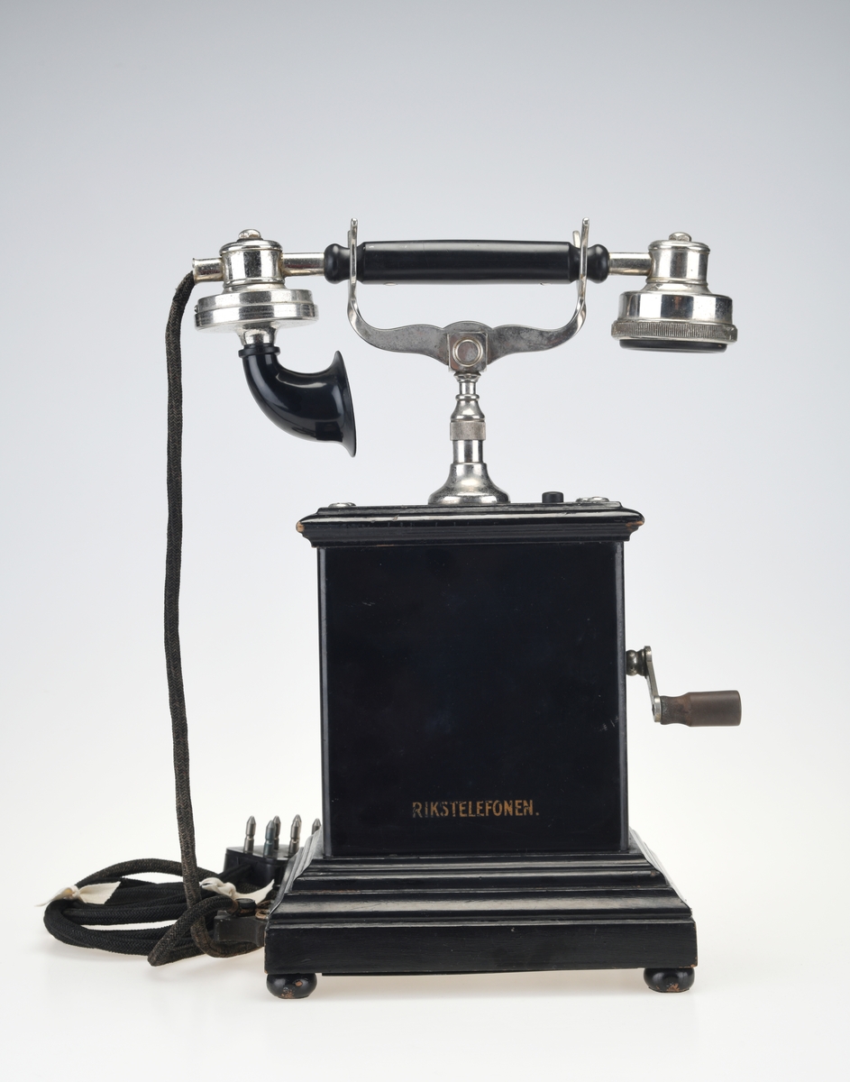 En bordtelefon av gammel modell laget av metall, tre og bakelitt. Foten på kassen har en ramme av svartlakkert metall og står på fire runde svartmalte trebein. På rammen er det montert på profilert treverk som også er svartmalt. Telefonkassen er av svart metall med en treplate med profilering på toppen som er skrudd fast. På midten er det en gaffel av sølvfarget metall der telefonrøret skal ligg. På den ene siden av kassen er det en sveiv av sølvfarget metall og håndtak av brun bakelitt. Ved siden av gaffelen er det en liten knapp som kan trykkes ned. Håndtaket på telefonrøret er av svart bakelitt. Høre- og snakkedelen er laget av blankt metall. På høredelen er det en rund bakelittplate med 7 hull. På snakkedelen er det et horn av svart bakelitt (har form som et lite grammofonhorn). Nede på den ene kortsiden går ledningen til telefonen ut. Den er lang og ender i et støpsel av svart bakelitt med seks metallplugger.