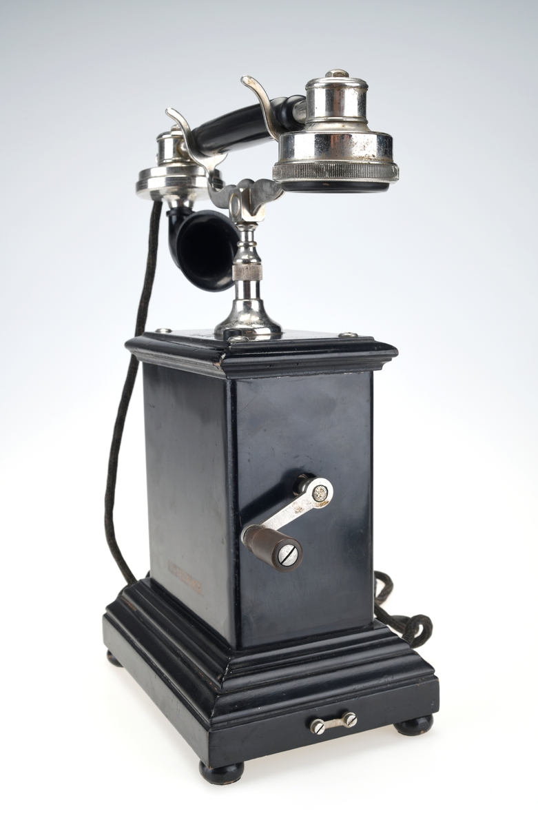 En bordtelefon av gammel modell laget av metall, tre og bakelitt. Foten på kassen har en ramme av svartlakkert metall og står på fire runde svartmalte trebein. På rammen er det montert på profilert treverk som også er svartmalt. Telefonkassen er av svart metall med en treplate med profilering på toppen som er skrudd fast. På midten er det en gaffel av sølvfarget metall der telefonrøret skal ligg. På den ene siden av kassen er det en sveiv av sølvfarget metall og håndtak av brun bakelitt. Ved siden av gaffelen er det en liten knapp som kan trykkes ned. Håndtaket på telefonrøret er av svart bakelitt. Høre- og snakkedelen er laget av blankt metall. På høredelen er det en rund bakelittplate med 7 hull. På snakkedelen er det et horn av svart bakelitt (har form som et lite grammofonhorn). Nede på den ene kortsiden går ledningen til telefonen ut. Den er lang og ender i et støpsel av svart bakelitt med seks metallplugger.