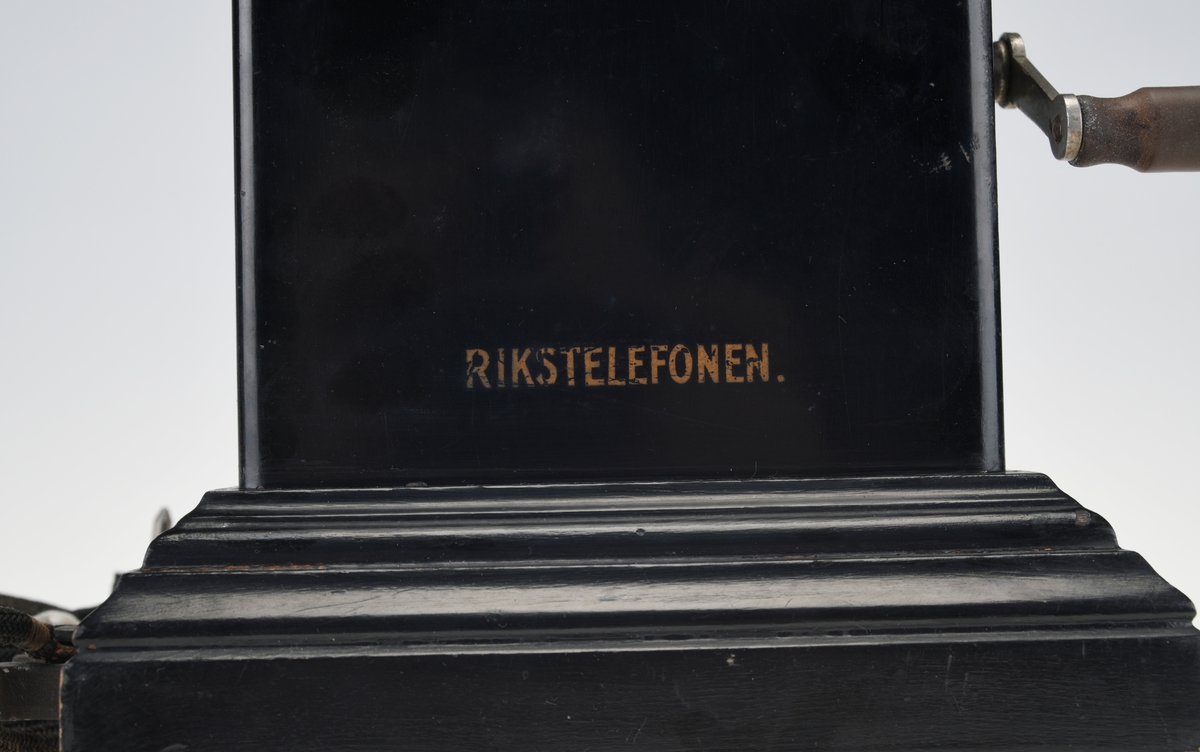 En bordtelefon av gammel modell laget av metall, tre og bakelitt. Foten på kassen har en ramme av svartlakkert metall og står på fire runde svartmalte trebein. På rammen er det montert på profilert treverk som også er svartmalt. Telefonkassen er av svart metall med en treplate med profilering på toppen som er skrudd fast. På midten er det en gaffel av sølvfarget metall der telefonrøret skal ligg. På den ene siden av kassen er det en sveiv av sølvfarget metall og håndtak av brun bakelitt. Ved siden av gaffelen er det en liten knapp som kan trykkes ned. Håndtaket på telefonrøret er av svart bakelitt. Høre- og snakkedelen er laget av blankt metall. På høredelen er det en rund bakelittplate med 7 hull. På snakkedelen er det et horn av svart bakelitt (har form som et lite grammofonhorn). Nede på den ene kortsiden går ledningen til telefonen ut. Den er lang og ender i et støpsel av svart bakelitt med seks metallplugger.