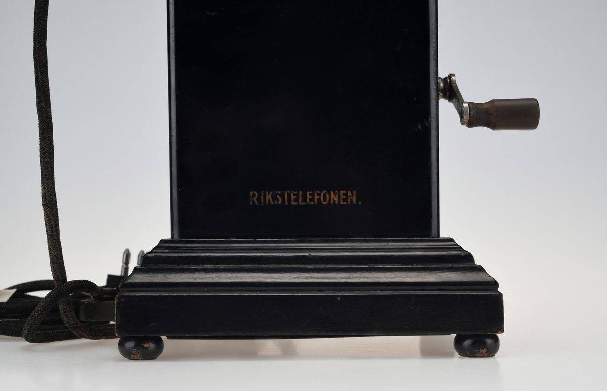 En bordtelefon av gammel modell laget av metall, tre og bakelitt. Foten på kassen har en ramme av svartlakkert metall og står på fire runde svartmalte trebein. På rammen er det montert på profilert treverk som også er svartmalt. Telefonkassen er av svart metall med en treplate med profilering på toppen som er skrudd fast. På midten er det en gaffel av sølvfarget metall der telefonrøret skal ligg. På den ene siden av kassen er det en sveiv av sølvfarget metall og håndtak av brun bakelitt. Ved siden av gaffelen er det en liten knapp som kan trykkes ned. Håndtaket på telefonrøret er av svart bakelitt. Høre- og snakkedelen er laget av blankt metall. På høredelen er det en rund bakelittplate med 7 hull. På snakkedelen er det et horn av svart bakelitt (har form som et lite grammofonhorn). Nede på den ene kortsiden går ledningen til telefonen ut. Den er lang og ender i et støpsel av svart bakelitt med seks metallplugger.