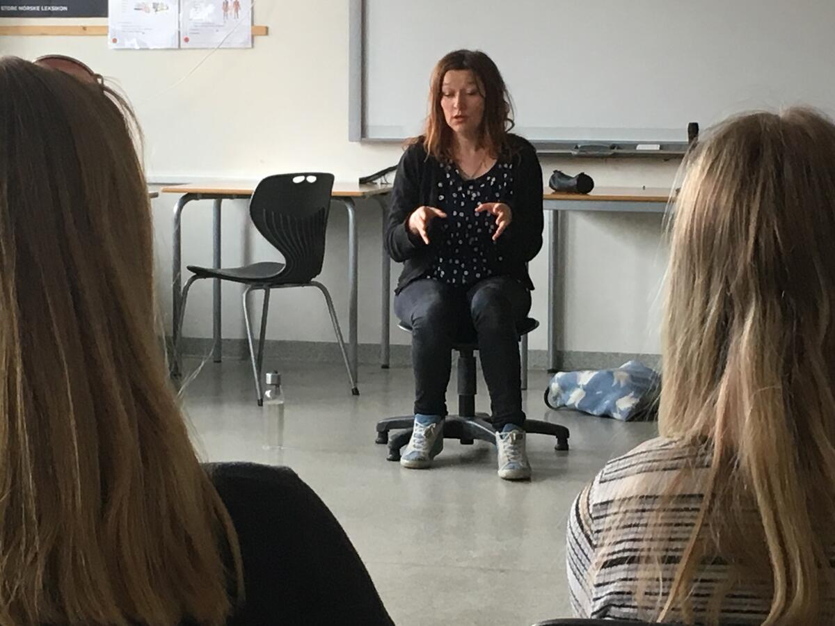 Vi ser ei kvinne som sitter på en stol foran i et klasserom, og to bakhoder med langt hår. Kvinnen gestikulerer med armene.