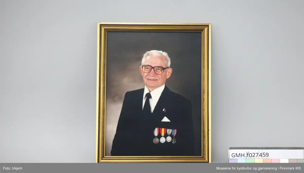 Studioportrett i farge av Hilmar Nordli som eldre mann. Nordli har vært disponent for Nord Norges Salgslag i Hammerfest. På bildet smiler han bredt, er kledd i svart dress og slips og hvit skjorte. Jakken er dekorert med fire utmerkelser, en av dem "Deltagermedaljen 9. april 1940–8. mai 1945", samt et merke.