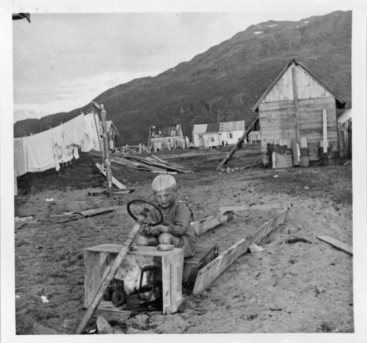 En ung gutt sitter på en hjemmelaget lastebil på stranda i Sørkjosen, trolig ved Nessevegen. Bildet er trolig tatt i 1946 og viser noe av bebyggelsen som stod igjen på neset etter 2. verdenskrig.