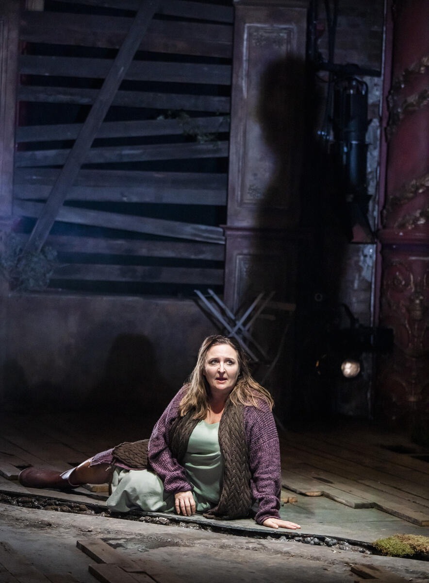 En teaterscene: En kvinne sitter på bakken i rufsete omgivelser.  