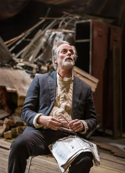 En teaterscene: En mann sitter på en stol med avisen i faget og kikker opp i luften