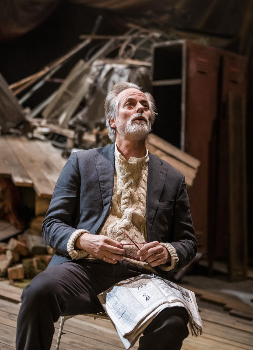 En teaterscene: En mann sitter på en stol med avisen i faget og kikker opp i luften