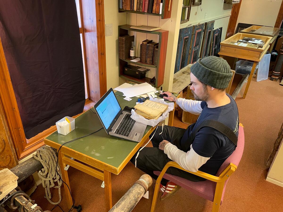 En mann som sitter i en rød stol i et rotete rom, foran seg har han en bok og en laptop på en grønn pult