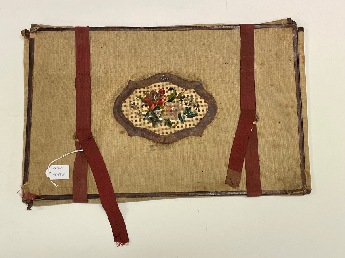 Av groft linne, kantat med brunt skinn, rödbruna bomullsband med spännen att binda det samman. I centrum en broderad utsmyckning föreställande blommor och blad.