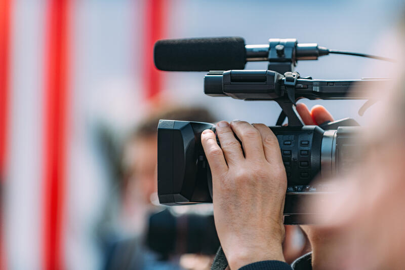 En hånd som holder et filmkamera med mikrofon, et ansikt er så vidt synlig i høre ende av bilde, men er ikke i fokus. I bakgrunnen ser man en person og noen røde striper, også ufokusert