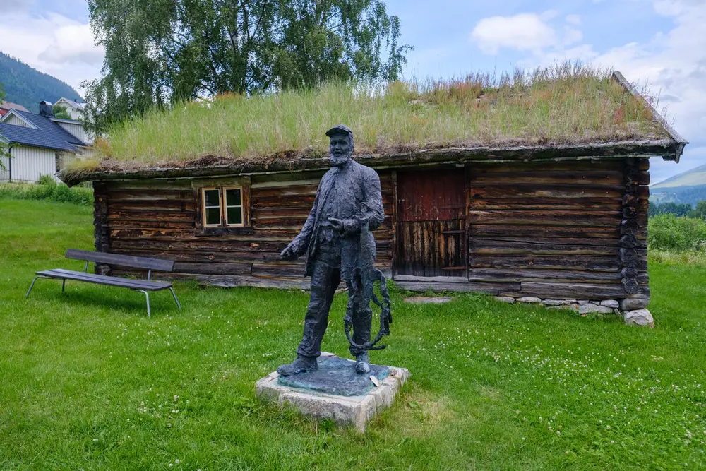 En mørk kobberstatue av en mann i gammeldagse klær står foran en liten brun hytte med gress på taket